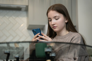 Teenage girl is using smartphone while sitting at glass table in kitchen with cup of coffee or tea, browsing internet, reading news, chatting in social networks, or playing mobile games