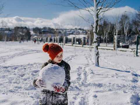 Girl in a winter duffel coat, bobble hat and gloves standing in a park carrying a heavy snowball to make a snowman, Ireland