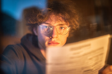 Young person reading a newspaper with focused expression in a dimly lit room during evening hours