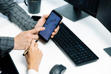 Cropped pointing hand with pen in businessman, checking with holding smartphone by woman investor online website program application with dynamic stock market at home office at back side. Infobahn.