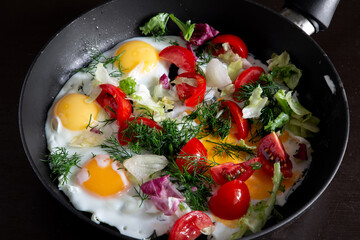 Fried eggs with vegetables and herbs in a frying pan on a black table