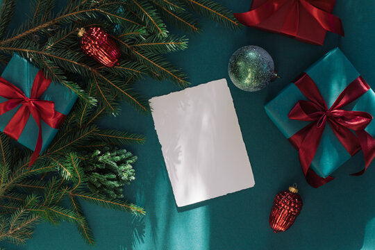Overhead view of a blank deckle edge card with wrapped gift boxes, festive Christmas ornaments and fir branches on a green background