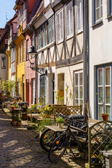 A narrow cobblestone street in Lubeck Germany with a row of houses and a church in the background. The street is lined with potted plants and outdoor seating, creating a cozy and inviting atmosphere