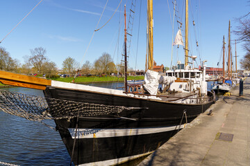 A large boat is docked at the pier in Lubeck Germany. The boat is black and white with a white stripe. The boat is surrounded by water and has a white flag on top