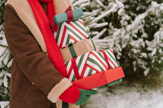 Close-up of a woman in a warm coat and gloves standing by snow covered Christmas fir trees holding a stack of wrapped Christmas gifts
