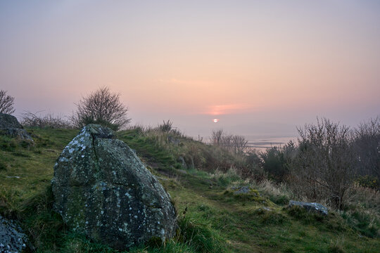 A tranquil sunset glows over a rugged coastline near Cramond Island, Edinburgh. Mossy rocks and low shrubs frame the calm sea and distant horizon.