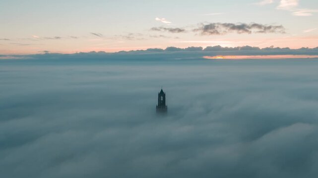Cinematic aerial video of church tower rising from low passing clouds and fog during sunrise in the city of Utrecht, the Netherlands