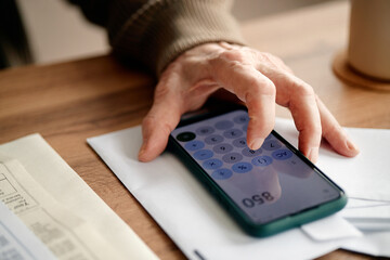 Senior woman analyzing invoices using smartphone calculator, hand pressing buttons on device while reviewing bills on wooden desk, focusing on financial calculations