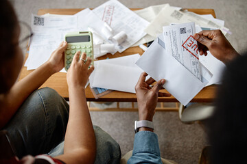 Two adult people, one woman and one Black man, analyzing utility bills and invoices at table, using calculator and opening envelopes, focusing on financial paperwork