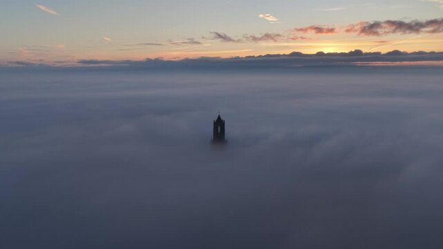 Cinematic aerial video of church tower rising from low passing clouds and fog during sunrise in the city of Utrecht, the Netherlands
