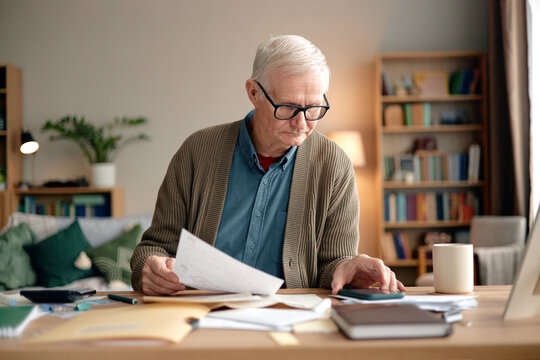 Senior man analyzing invoices and bills at desk, holding documents in one hand while using calculator with other hand, surrounded by paperwork and laptop in home office