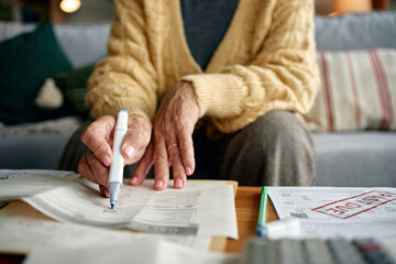 Senior woman reviewing invoices and bills at home, holding pen and analyzing financial documents on...