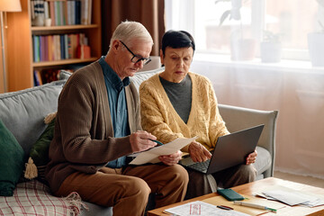 Senior man and senior woman sitting on sofa analyzing invoices and bills together, man holding documents and woman using laptop, financial paperwork on table