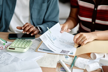 Woman and Black man analyzing invoices and bills at desk, using calculator and smartphone, reviewing financial documents together, hands holding paperwork and receipts