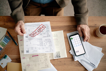 Senior man analyzing overdue invoices and bills at desk, holding past due notice in hand, using smartphone calculator for financial calculations, surrounded by paperwork