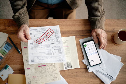 Senior man analyzing overdue invoices and bills at desk, holding past due notice in hand, using smartphone calculator for financial calculations, surrounded by paperwork
