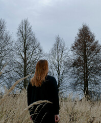 A woman with long brown hair stands alone in a golden meadow, surrounded by dry grass and trees. She gazes into the distance, embodying tranquility in nature.