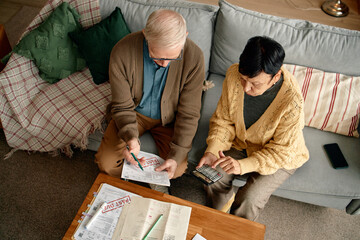 Senior man and senior Asian woman sitting on sofa reviewing invoices and bills together, using calculator and pen, analyzing financial documents on wooden table