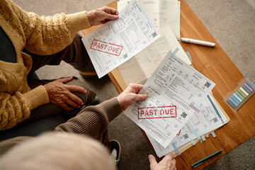 Senior man and senior woman reviewing overdue invoices and bills at table, holding documents marked past due, analyzing financial paperwork together