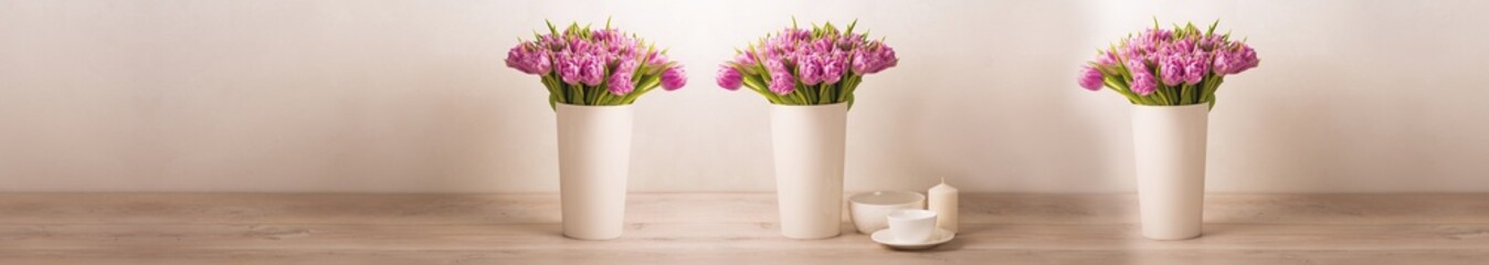 Elegant interior composition with pink tulips in white vases and candles on wooden table against soft beige wall background
