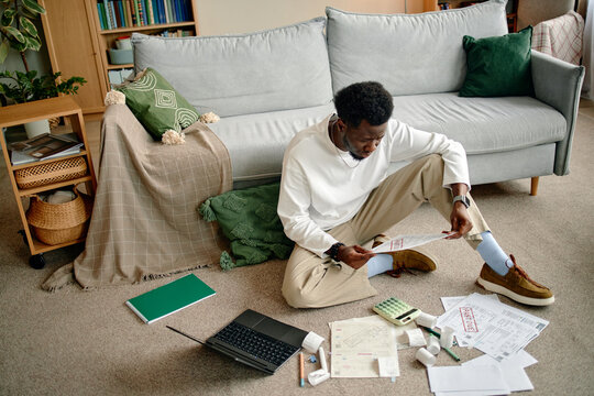 Black young man sitting on floor analyzing invoices and bills with laptop, calculator, documents and receipts spread around, focused on financial paperwork in living room