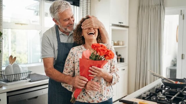 Senior husband surprising his joyful wife in the kitchen with a bouquet of fresh roses.