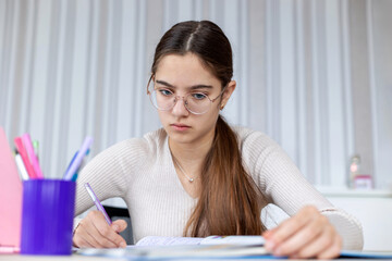 Teen girl studying and writing in notebook. A focused teenage girl wearing glasses writes notes in a notebook while studying at a desk with pens and textbooks.