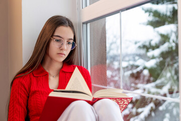 Teen girl reading book by snowy window. A young girl in a red sweater and glasses reads a book while sitting by a window with a snowy winter scene outside.