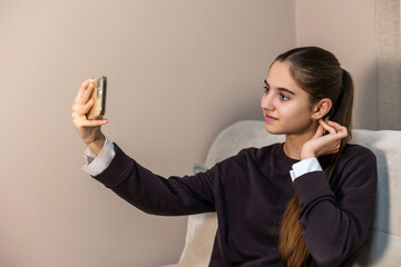 Teen girl taking a selfie and adjusting earring. Young girl poses for a selfie with her smartphone while fixing her earring, sitting comfortably against a beige wall indoors.