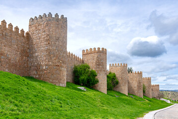 A portion of Ávila’s ancient stone walls, with tall rounded towers rising above a bright green embankment and set beneath a softly clouded sky that creates a calm atmosphere