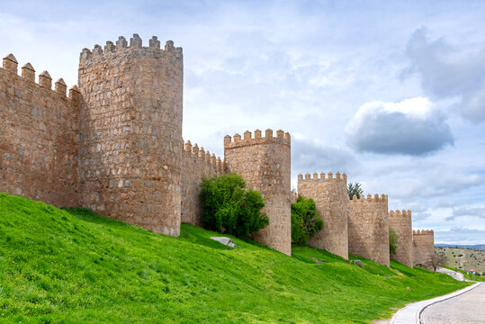 A portion of &Aacute;vila&rsquo;s ancient stone walls, with tall rounded towers rising above a bright green embankment and set beneath a softly clouded sky that creates a calm atmosphere