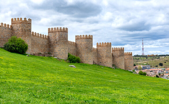 A portion of &Aacute;vila&rsquo;s ancient stone walls, with tall rounded towers rising above a bright green embankment and set beneath a softly clouded sky that creates a calm atmosphere