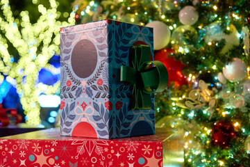 A patterned gift box with a green ribbon sits on a red present beside a glowing tree. Warm white fairy lights and ornaments sparkle in the background.