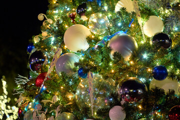 Shiny baubles and white ornaments fill the evergreen branches. Warm LED lights sparkle and glow in the festive night scene.