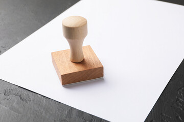 One wooden stamp and blank sheet of paper on dark textured table, closeup. Space for text