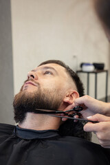 Barber shaving bearded customer in modern style barbershop. Male Beard hair cutting with scissors close up. Man client visit gentleman grooming beauty salon for evens the beard