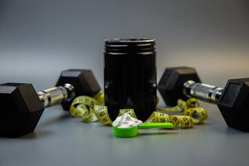 Black protein powder container is placed with dumbbells and measuring tape on gray surface, highlighting fitness, health, and nutrition essentials for workout enthusiasts