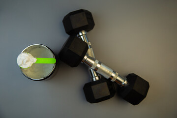 Black weights and a protein shake with a green spoon are arranged on a gray background, symbolizing fitness, strength training, and healthy nutrition choices