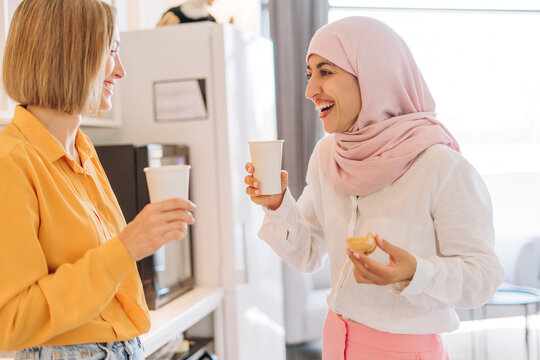 Two happy businesswomen wearing stylish casual clothes, one female hijab talking having coffee break