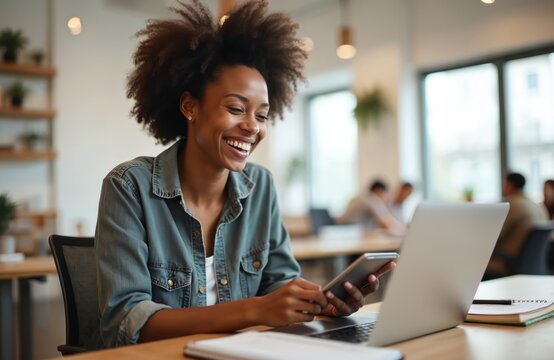Happy afro woman works in modern co-working space using laptop, smartphone. Smiling female freelancer enjoys online project. Business lady uses wireless tech device in office for remote work,
