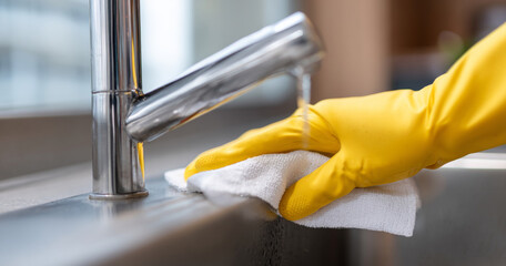 Close-up of hand wearing yellow rubber glove cleaning stainless steel kitchen sink with white cloth under running water faucet