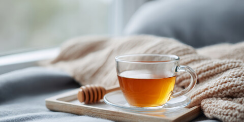 Cozy morning scene with a glass cup of tea on a wooden tray next to a honey dipper and soft knitted blanket by the window