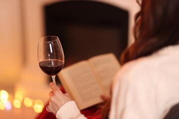 Woman with red wine reading book near fireplace at home, closeup