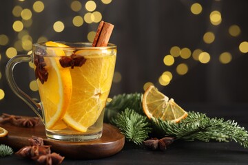 Delicious mulled wine with slices of orange, spices and fir branches on table against black background with blurred Christmas lights, closeup. Bokeh effect