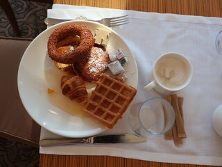 A white plate showcases a sesame bagel ring, a waffle, a croissant, and assorted pastries with sugar cubes, paired with a coffee cup on a light, stylish table setting.