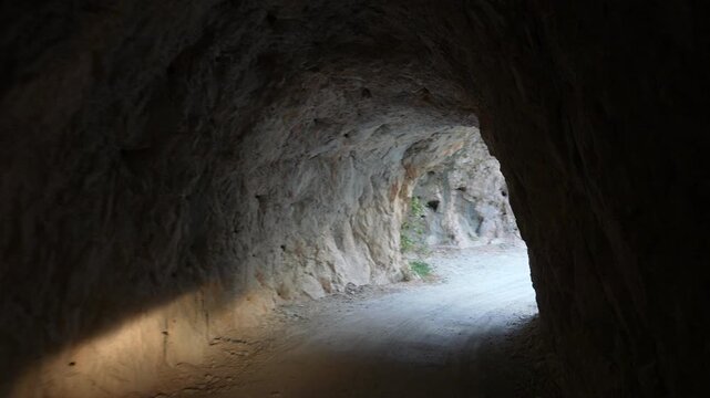 A rocky tunnel on a narrow canyon road in Kemaliye shifts from darkness to bright daylight. The rough stone walls, dust and curved mountain path create a dramatic and cinematic transition scene.