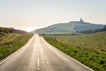 Straight stretch of a  coastal road at sunset in spring. A clifftop lighthouse is visible in distance. Seven Sistes cliffs, England, UK.