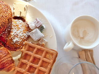 A hearty morning scene featuring waffles, croissant, sesame bun, and assorted confections alongside a creamy cup of coffee on a bright white table.