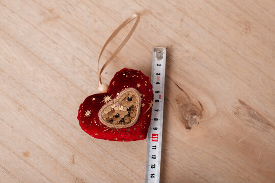 Top view of a red sequin Christmas heart ornament next to a measuring tape on a wooden table, highlighting its dimensions for product listing, holiday advertising, and festive decoration concepts.