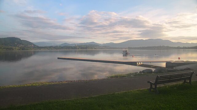 Lake Comabbio at Corgeno town, Italy. Peace and tranquility at dawn with a bench in the foreground. In the background the towns of Varano Borghi, Ternate, Comabbio and the Campo dei Fiori massif 
